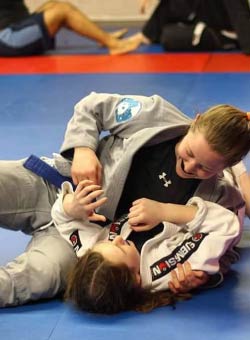 Coach Lesley Steiner works with a female youth Brazilian Jiu-Jistu student from side-contol in BJJ classes. Both Coach Steiner and the student are wearing white gis.