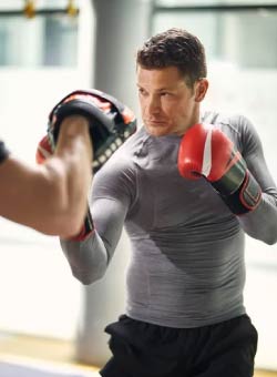 A Caucasian male wearing a gray rashguard and red boxing gloves punches black mitts in an adult kickboxing muay thai class at Excelsior MMA.