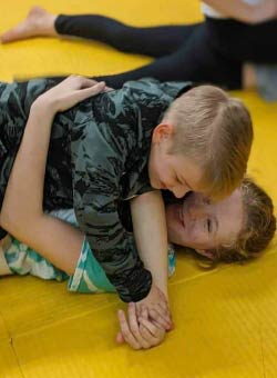 Two youth Brazilian Jiu-jitsu students grappling. The student on the top is wearing a camouflage top and is in the mount position. He has his opponent's right arm across his chest.